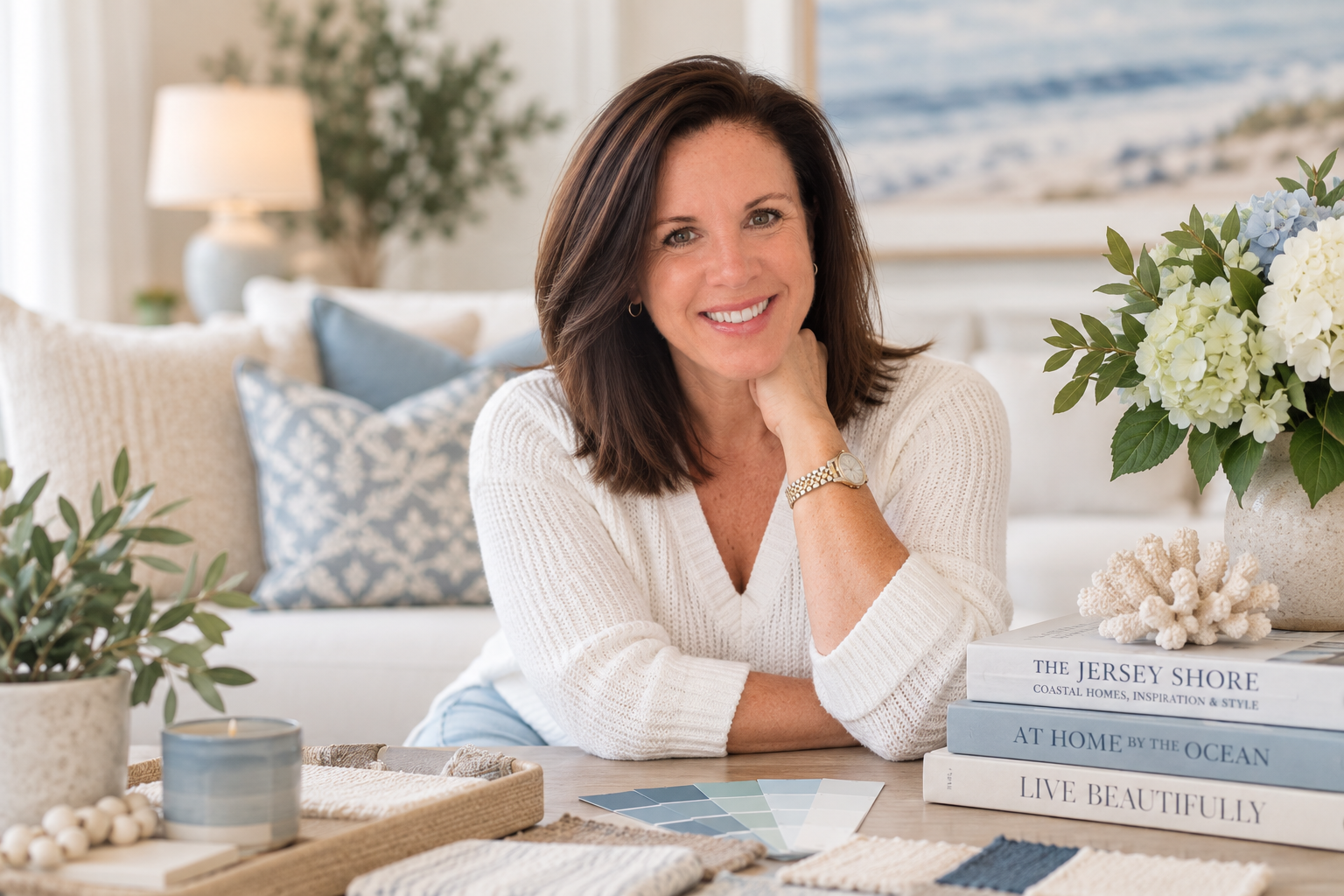 Jodi Kelly sitting at a coffee table with coastal blues, creams and sandy decor and furniture surrounding her.